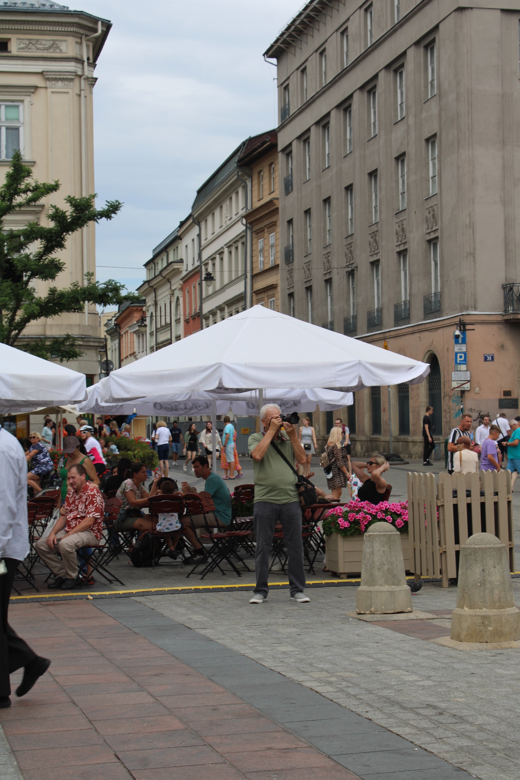 A man stands in front of some outdoor restaurant seating with a camera to his face