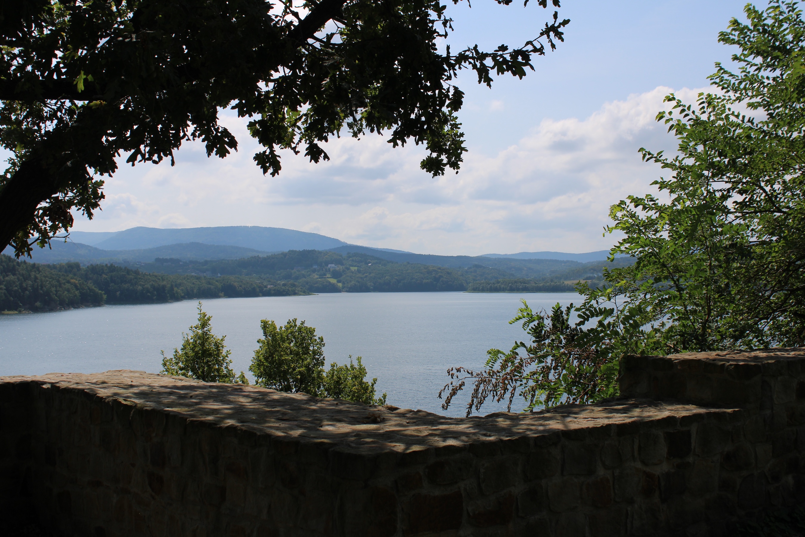 A view of huge lake with rolling hills in the back and a beautiful sky. A stone wall and trees frame the photo.