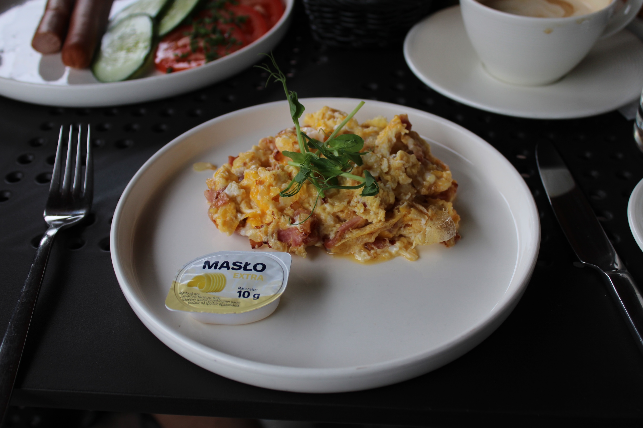 A white ceramic frisbee-looking plate contains a small portion of eggs, bacon and onions. There's some green-leaf garnish, and a small disposable container of butter as well.