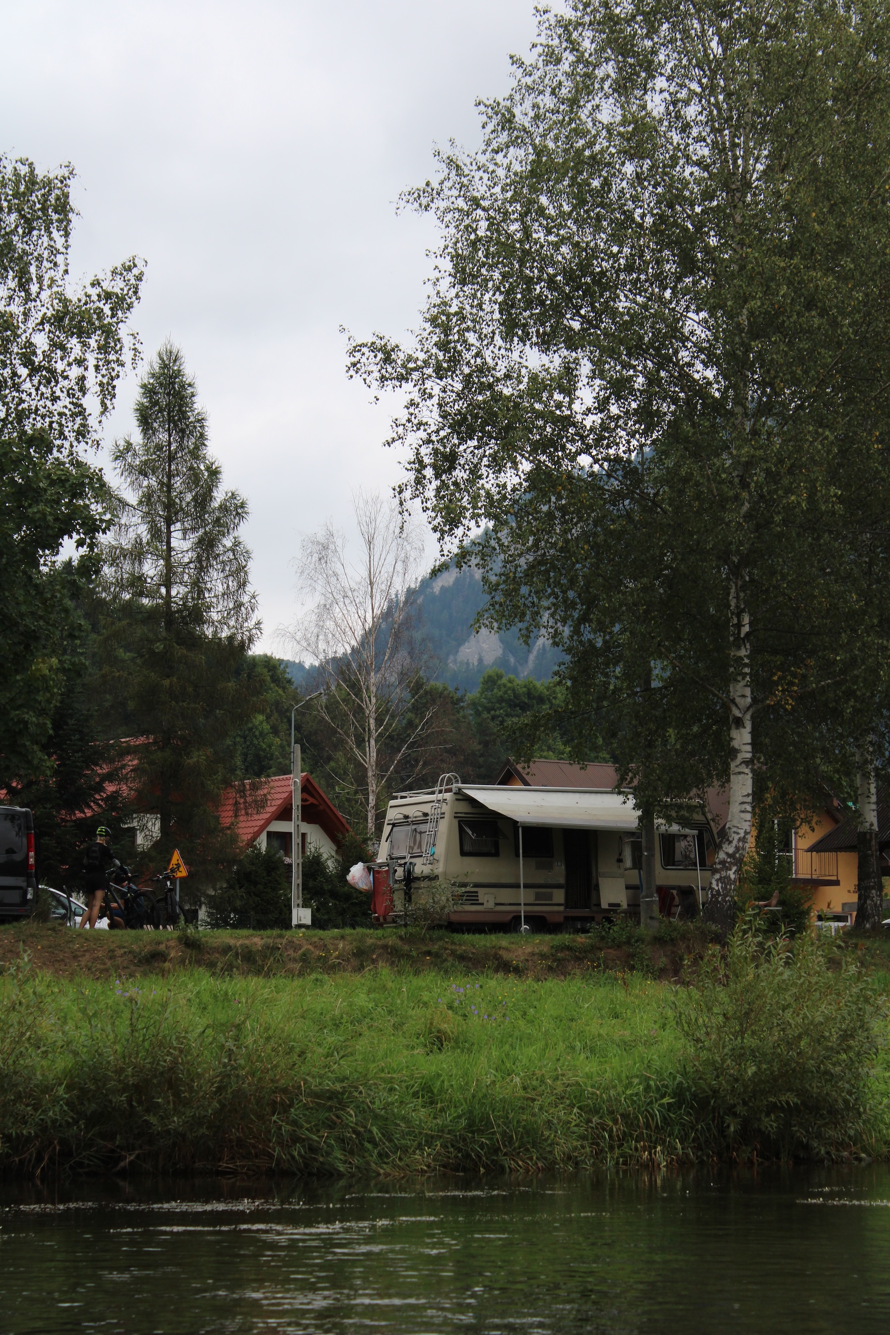 A camper van on the river edge. Lots of green grass and trees around it.