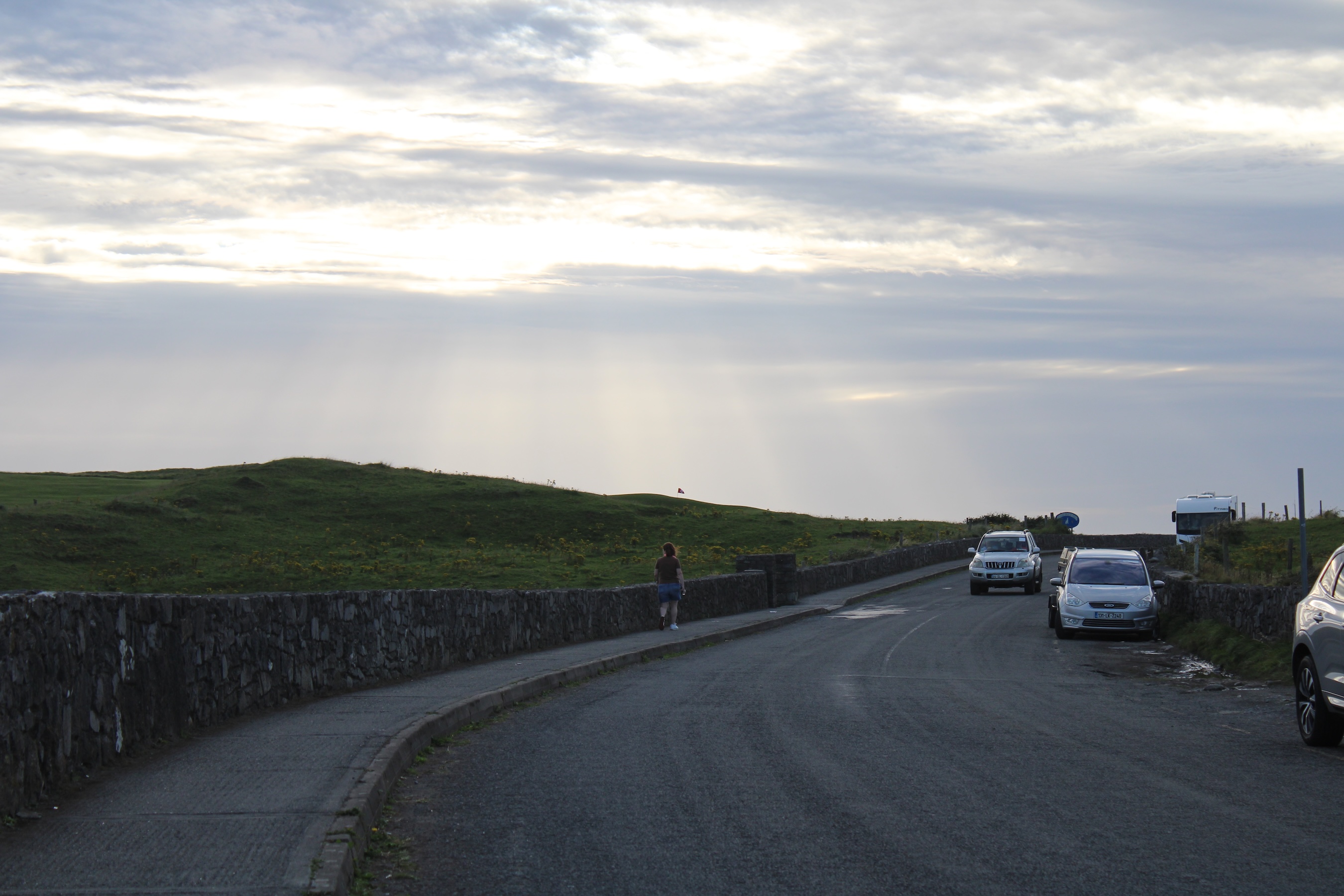 A roadway curves to the right. A golf course to the left is lit by a cloudy sky letting in god rays from above.